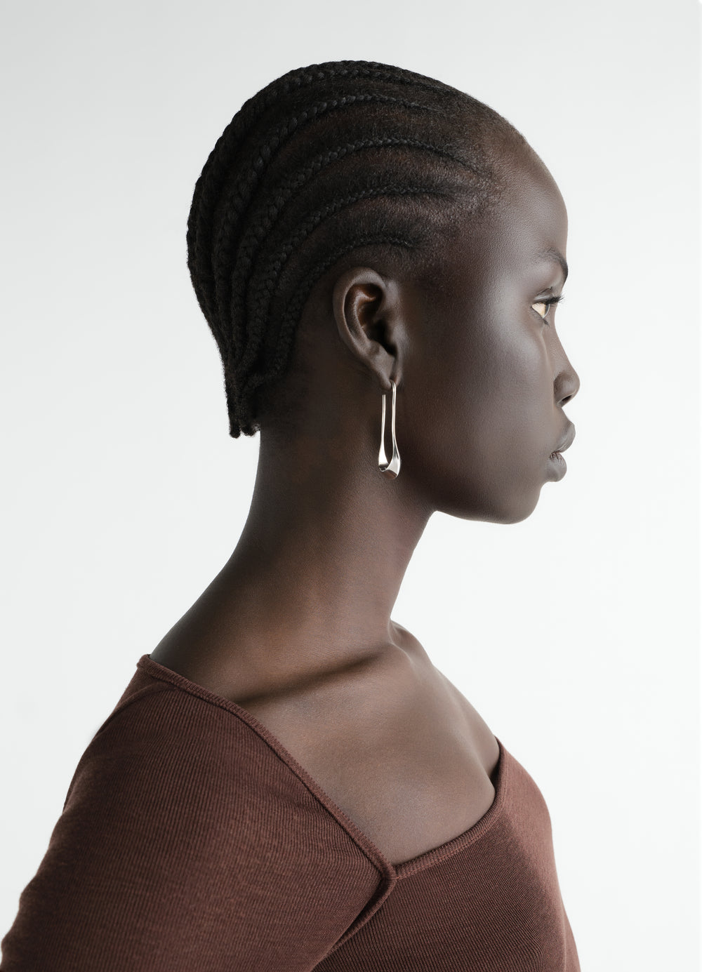 Short silver drop earrings by LEMAIRE worn by a model in profile with a brown top on white background