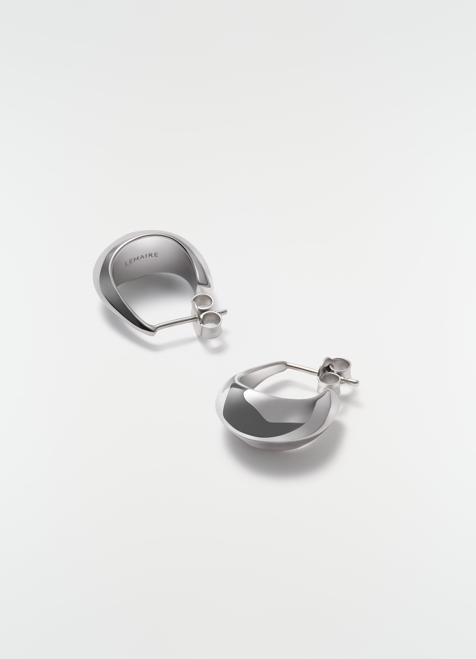 Curved mini drop earrings in polished silver by lemaire styled on plain white background close-up detail