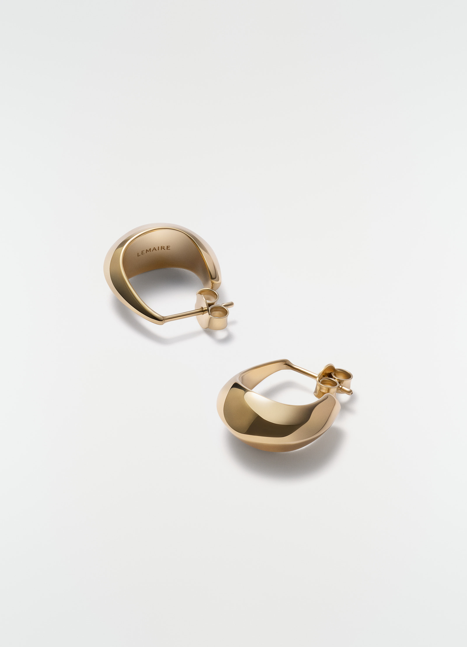 Curved mini drop earrings in gold vermeil metal by lemaire styled on plain white background close-up detail.