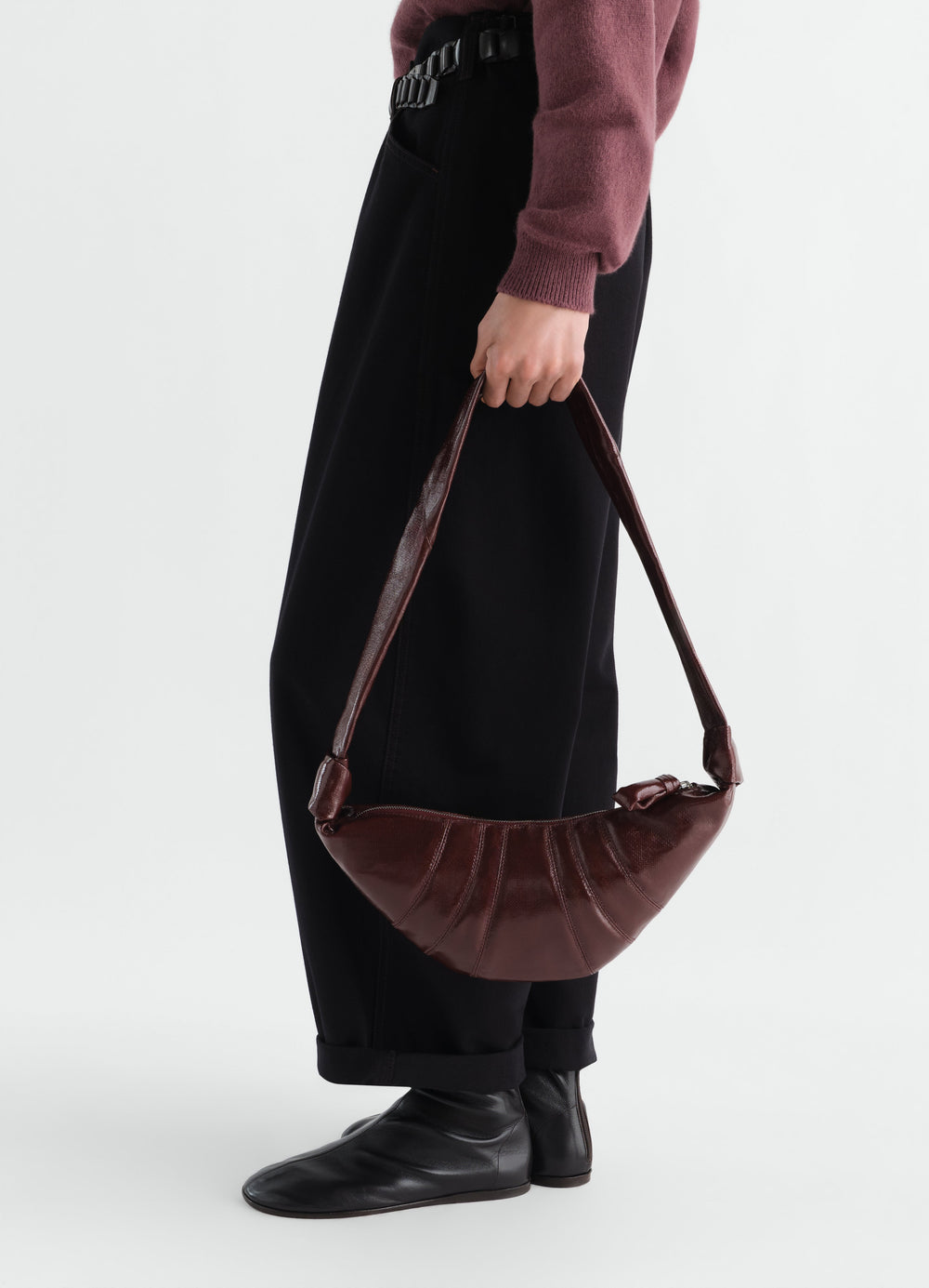 Medium croissant-shaped leather bag in dark brown LEMAIRE worn by model side view with black pants and boots