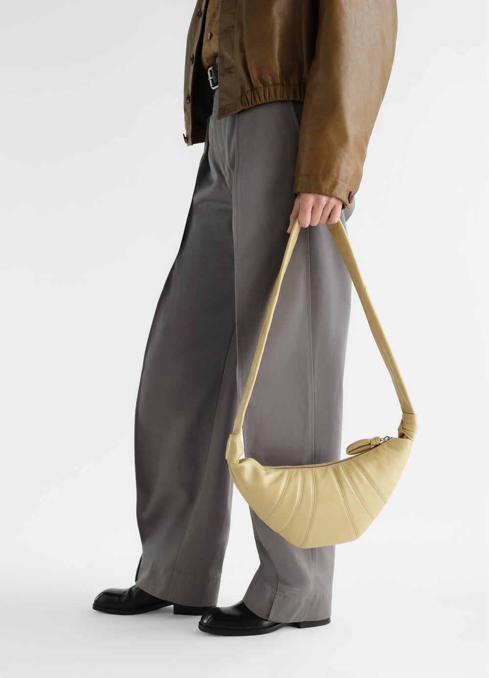 Small beige leather croissant bag by LEMAIRE worn by model with brown jacket and grey pants side view