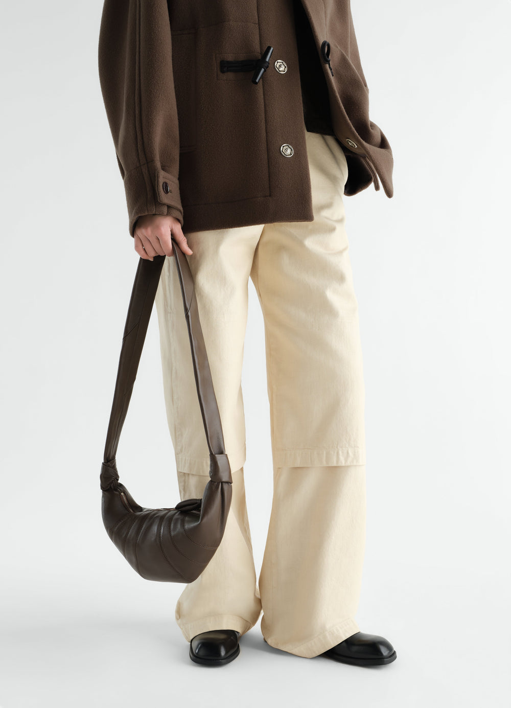 Small brown leather Croissant bag LEMAIRE held by model with cream trousers and brown jacket front view