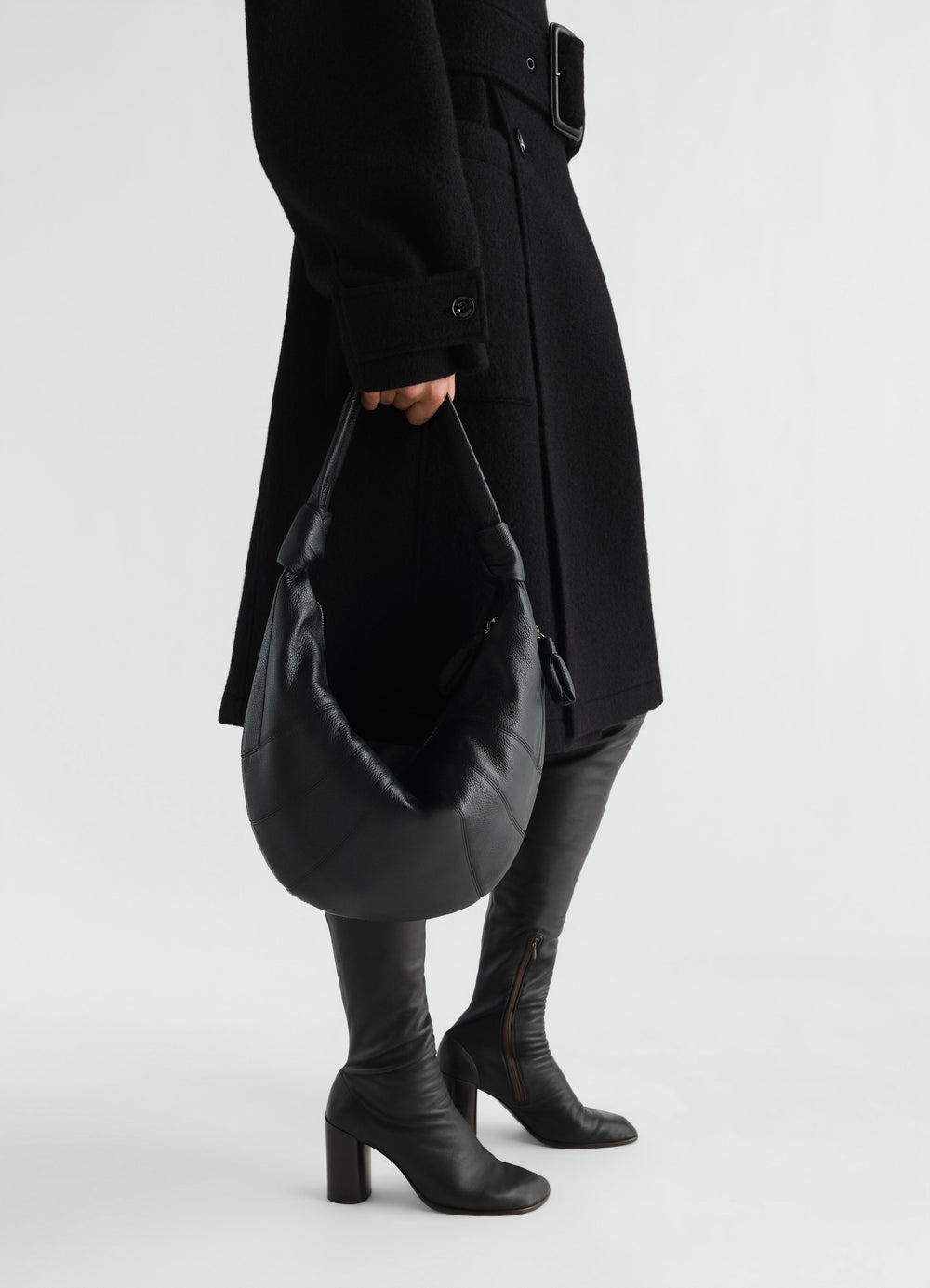 Fortune Croissant leather bag in black LEMAIRE worn by model holding bag with black coat and boots side view