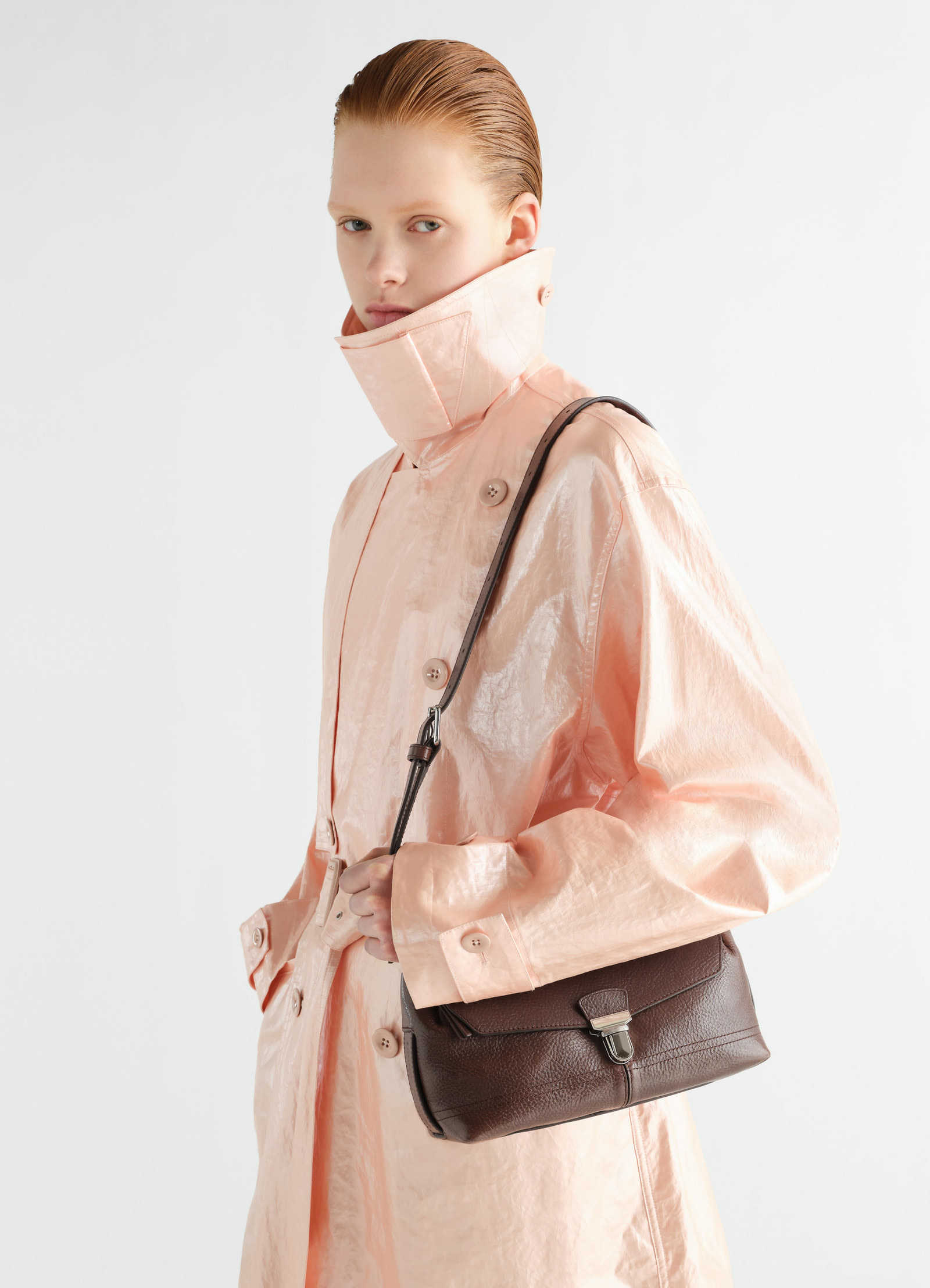 Small dark brown leather gear bag by lemaire worn crossbody by model in pale pink shiny coat front view