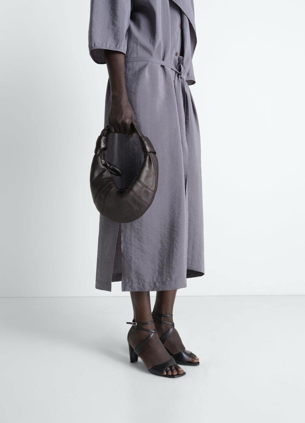 Mini Fortune Croissant bag in dark brown leather by LEMAIRE worn by model with lilac dress and black sandals side view
