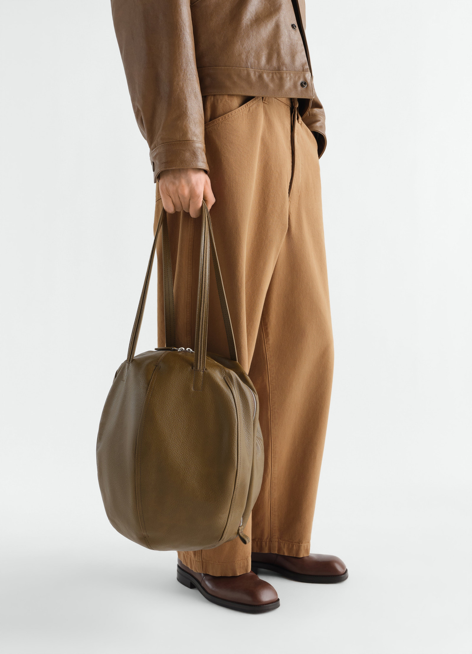 Hobo helmet bag in olive green grained leather lemaire worn by model side view with brown pants and leather jacket