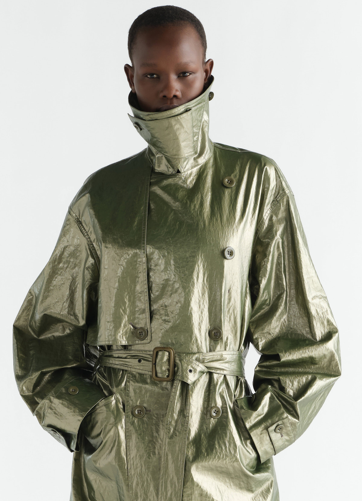 Trench coat in metallic green shiny coated linen with belt and high collar worn by model lemaire
