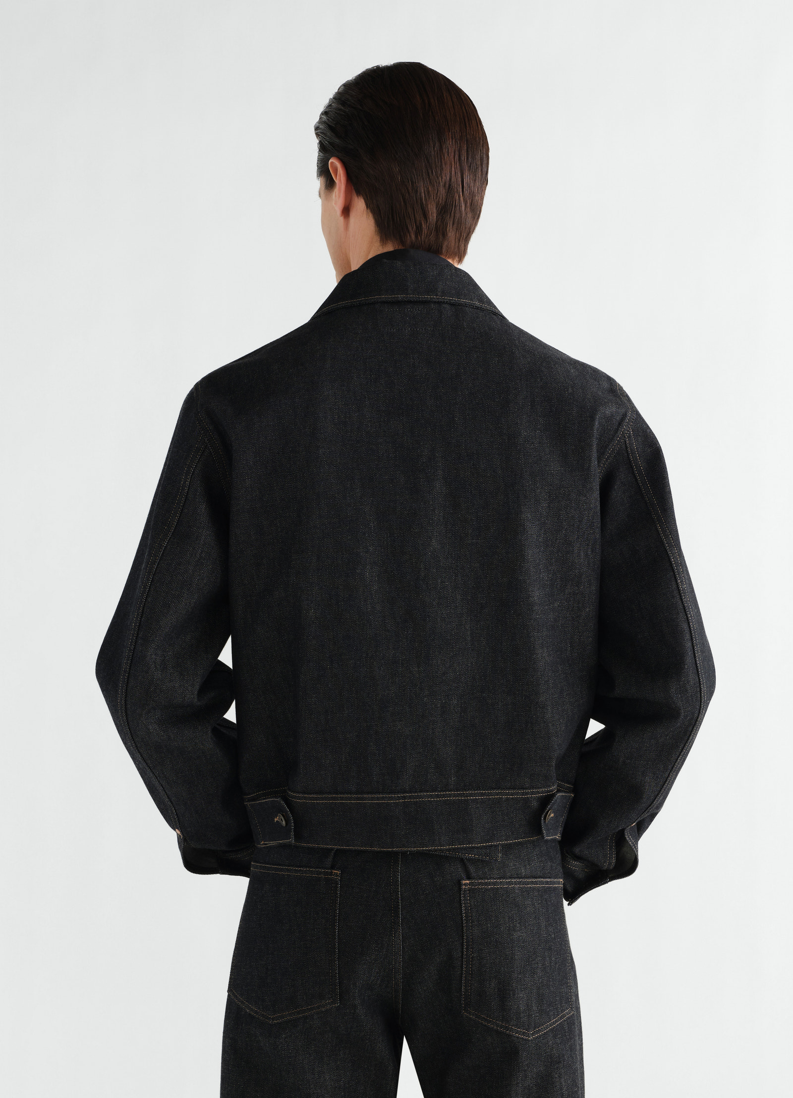 Trucker jacket in slate black denim with brown stitching by lemaire worn by a model back view