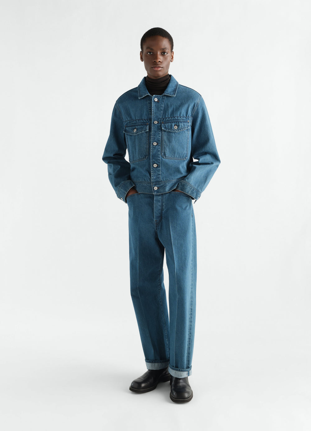 Trucker jacket in indigo selvedge denim by LEMAIRE worn by model with matching jeans on white background