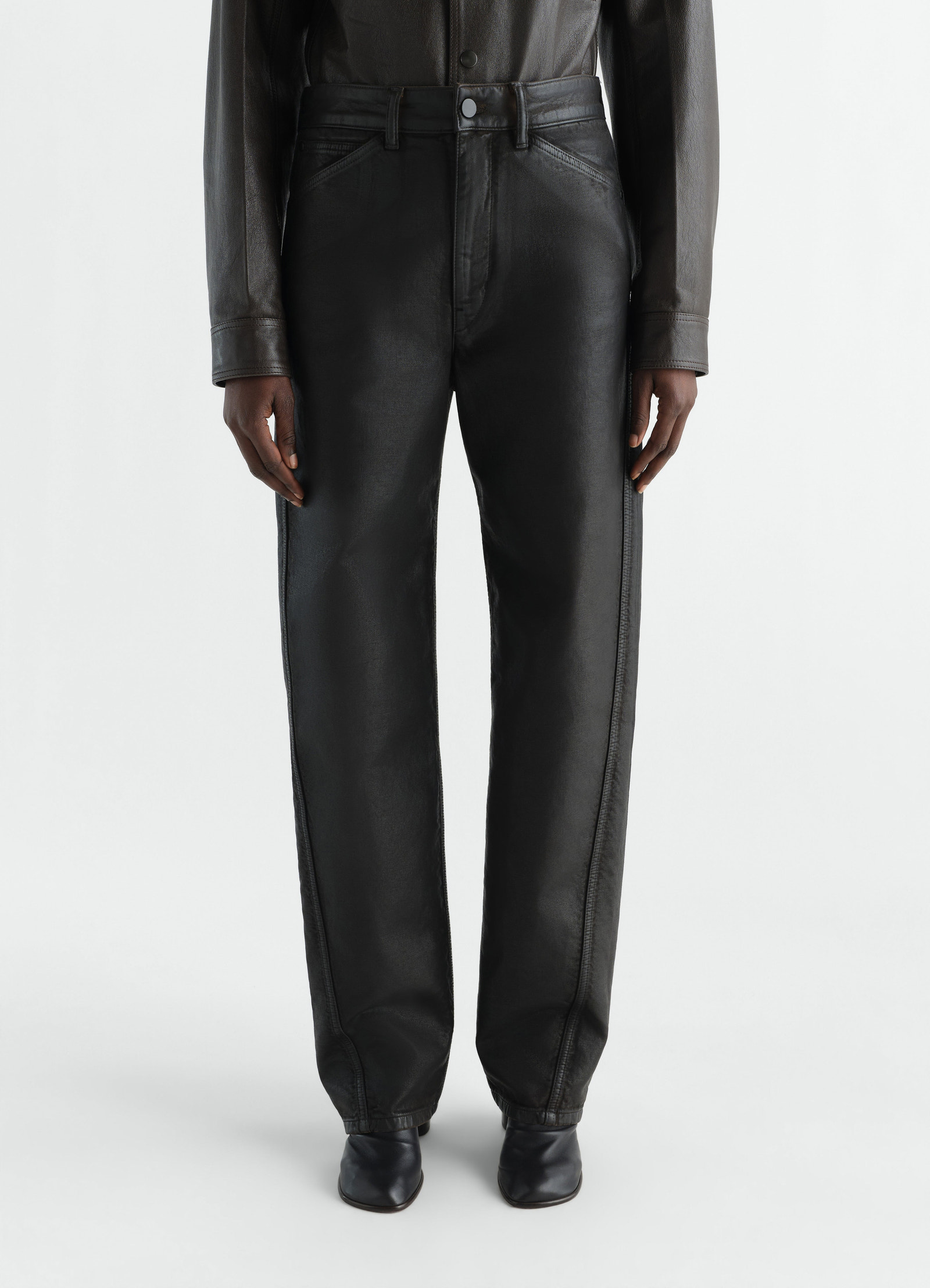 Twisted five-pocket black coated denim pants by lemaire worn by model front view with black leather boots