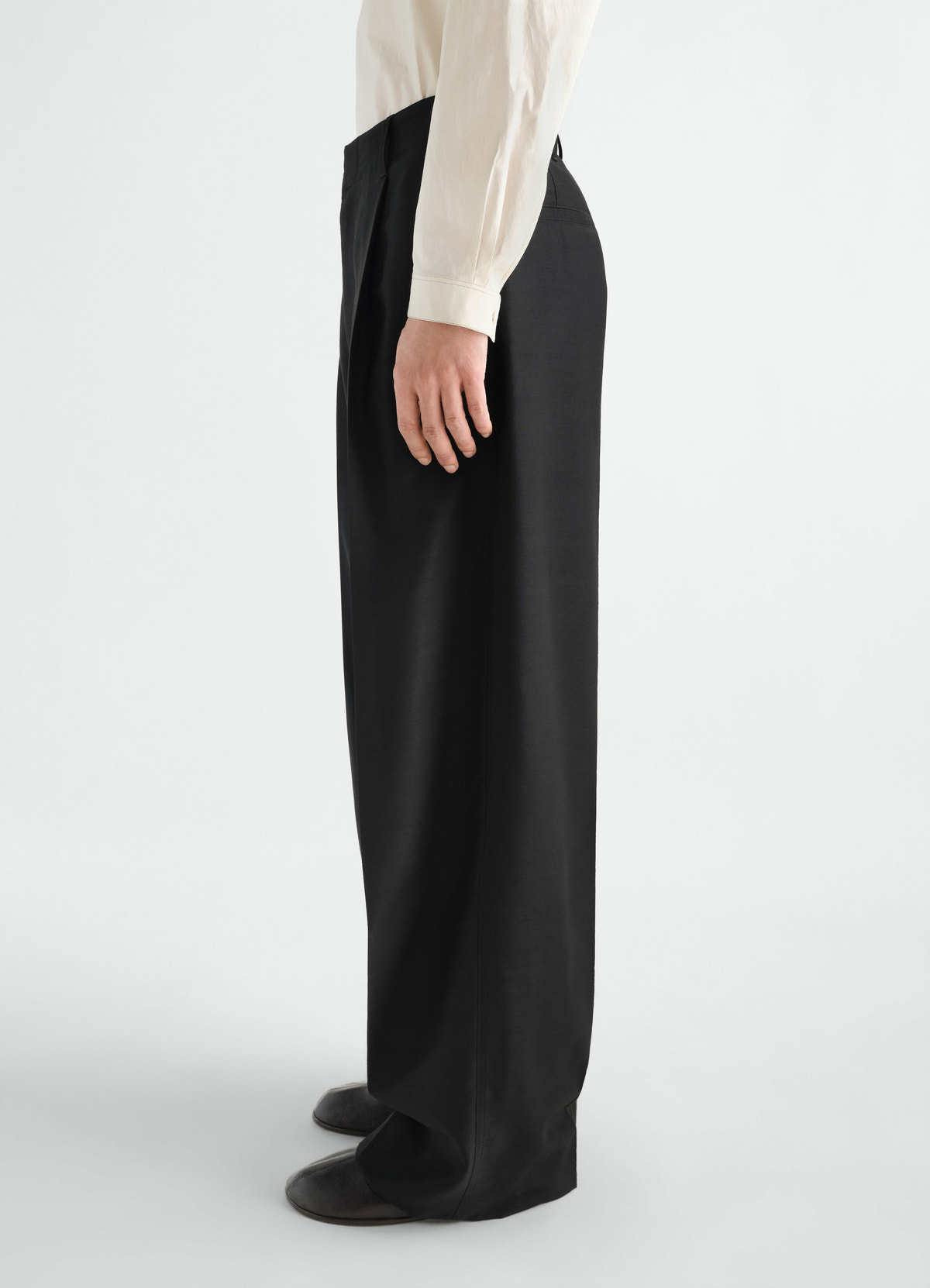 Single-pleat black wool mohair pants by lemaire worn by a model side view with cream shirt and black shoes
