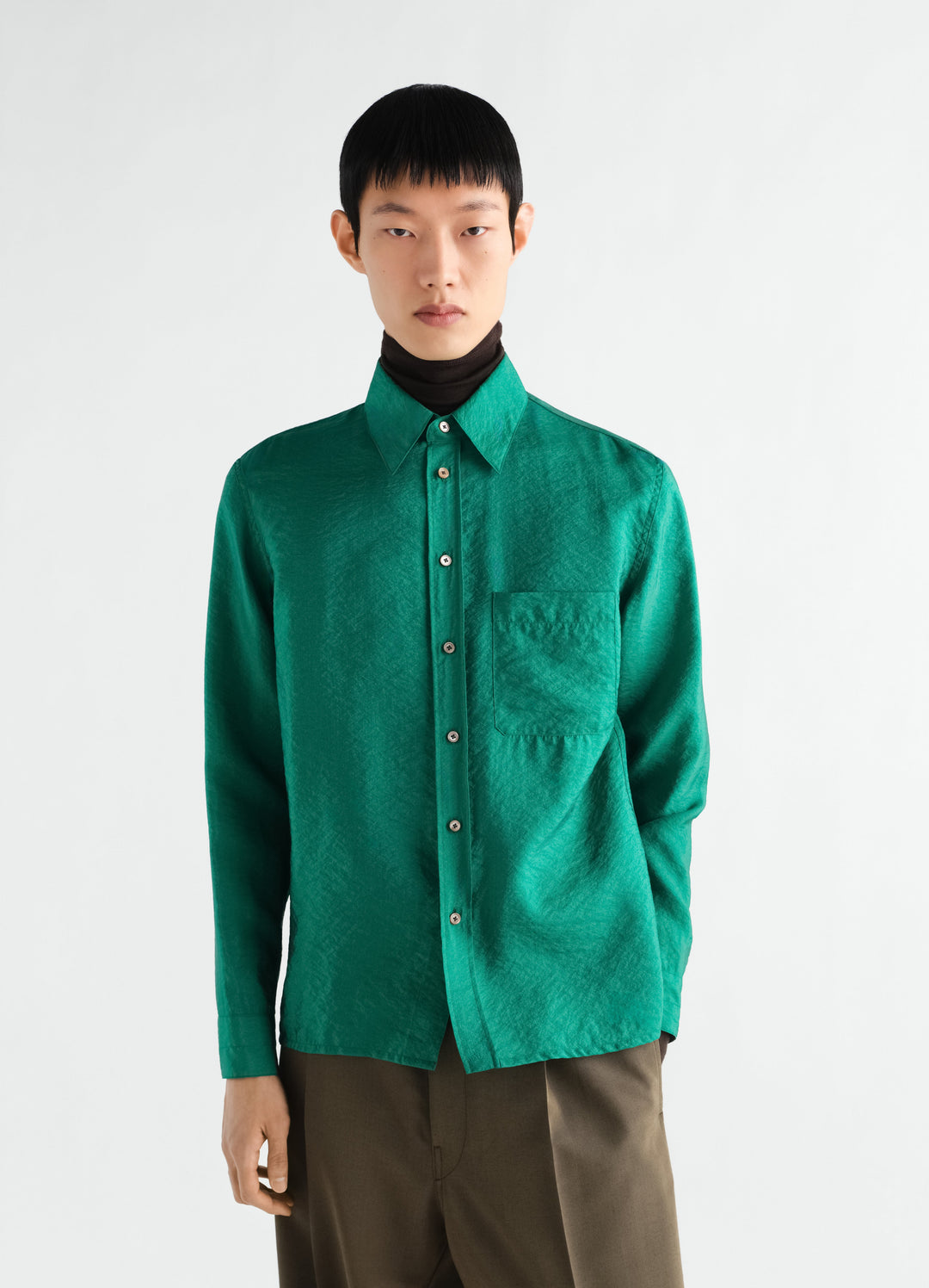 Fitted green shiny cupro shirt by LEMAIRE worn by model front view with black turtleneck and brown pants