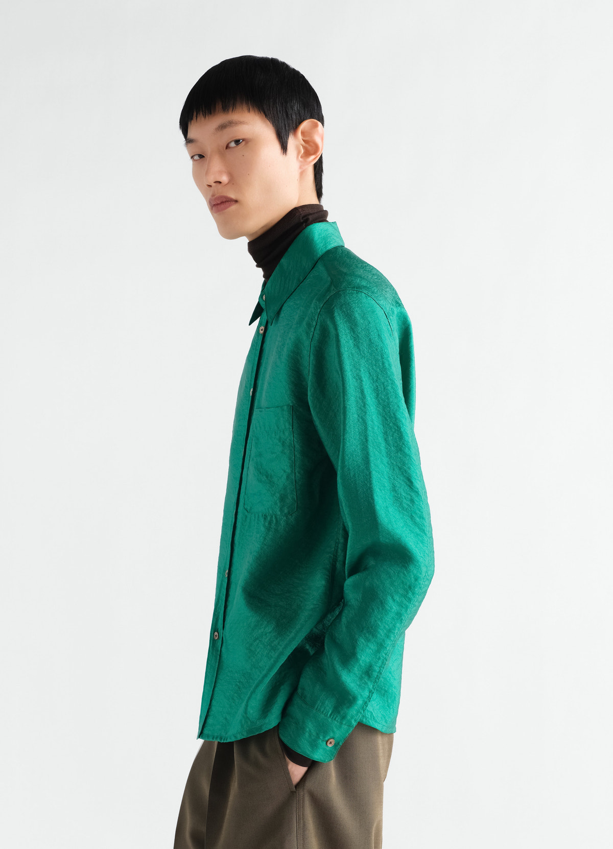 Fitted shiny green cupro shirt by lemaire worn by model side view with brown turtleneck and olive pants