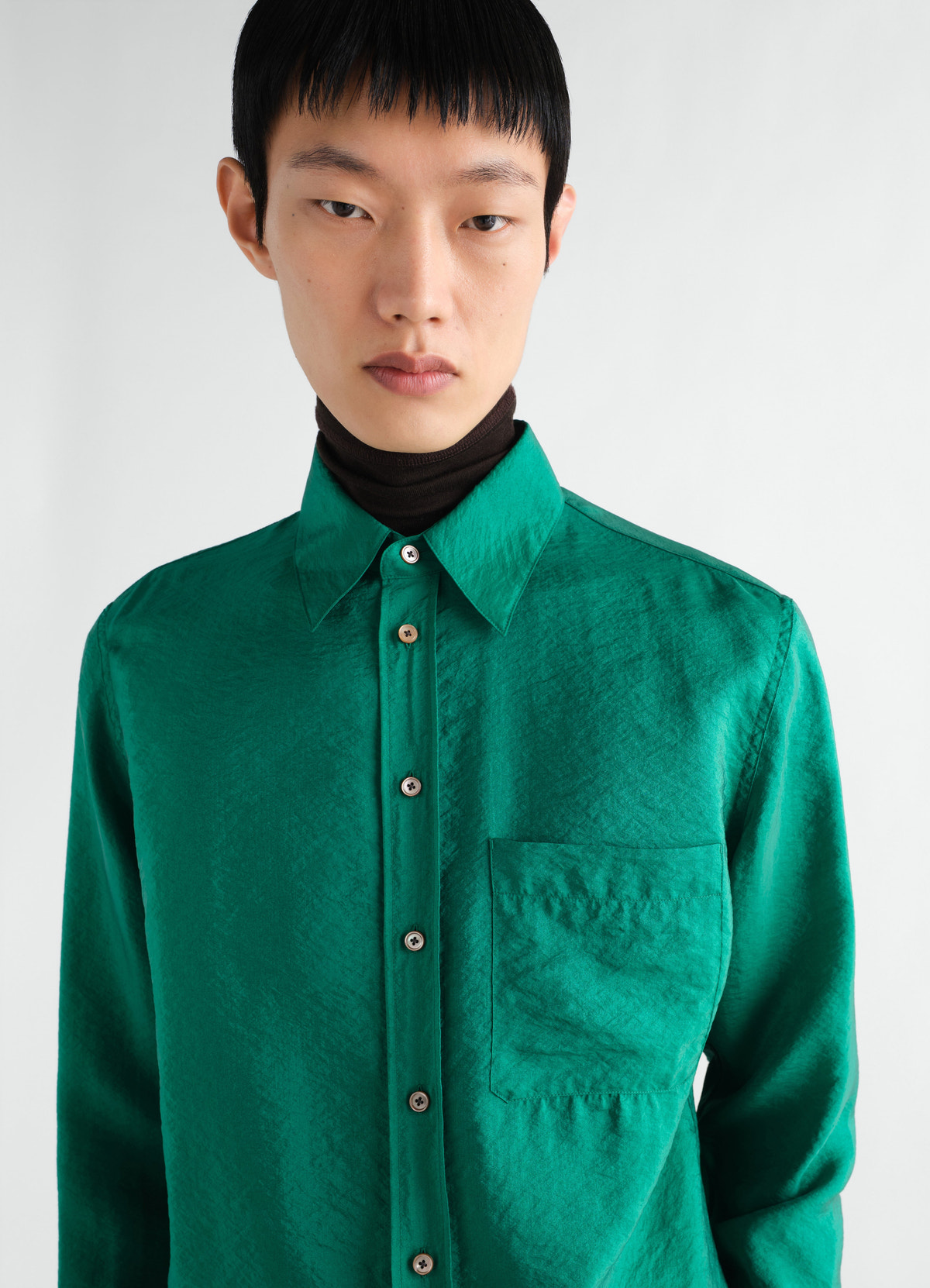 Fitted green shiny cupro shirt with pocket worn by model layered over black turtleneck lemaire front view