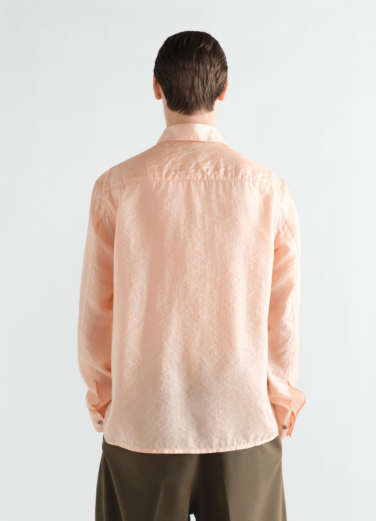 Fitted light peach shiny cupro shirt by lemaire worn by model back view with olive pants