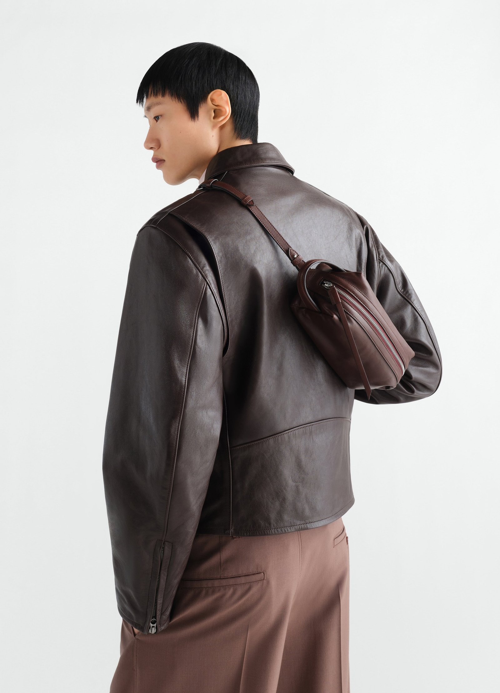 Small brown nappa lambskin care bag by lemaire worn crossbody on model back with leather jacket side view