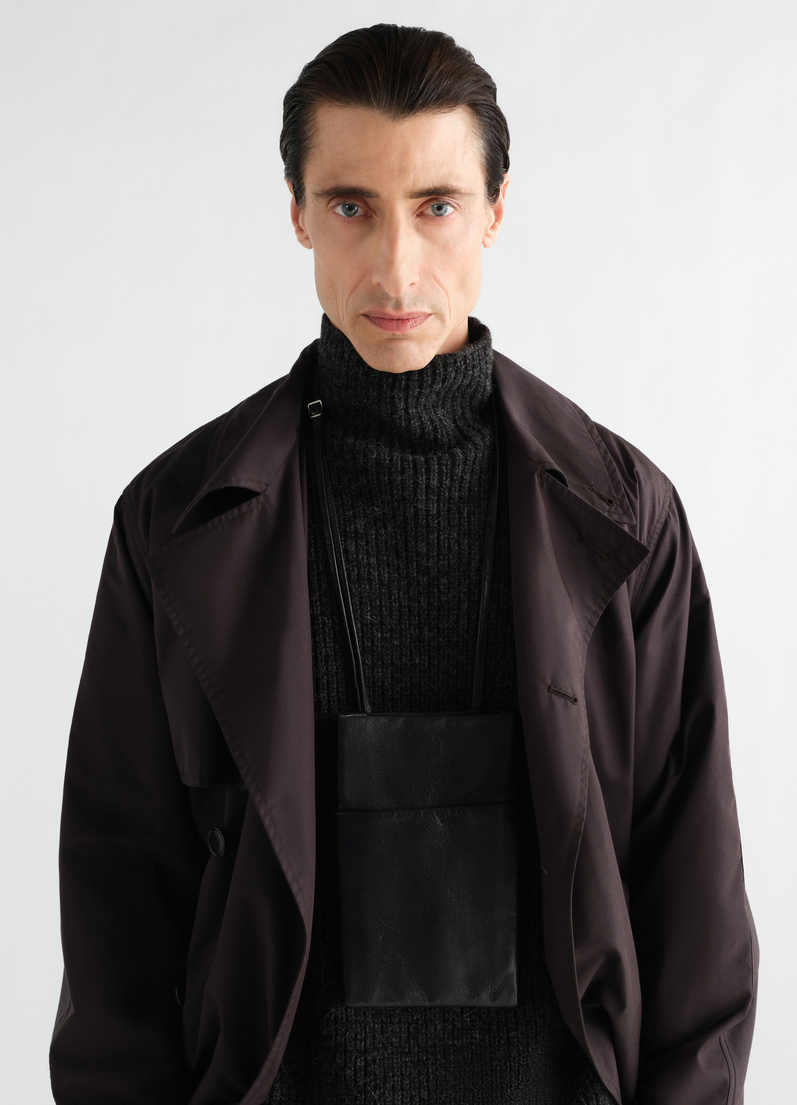 Book pouch in black grained leather worn by model with dark knit and oversized brown jacket lemaire front view