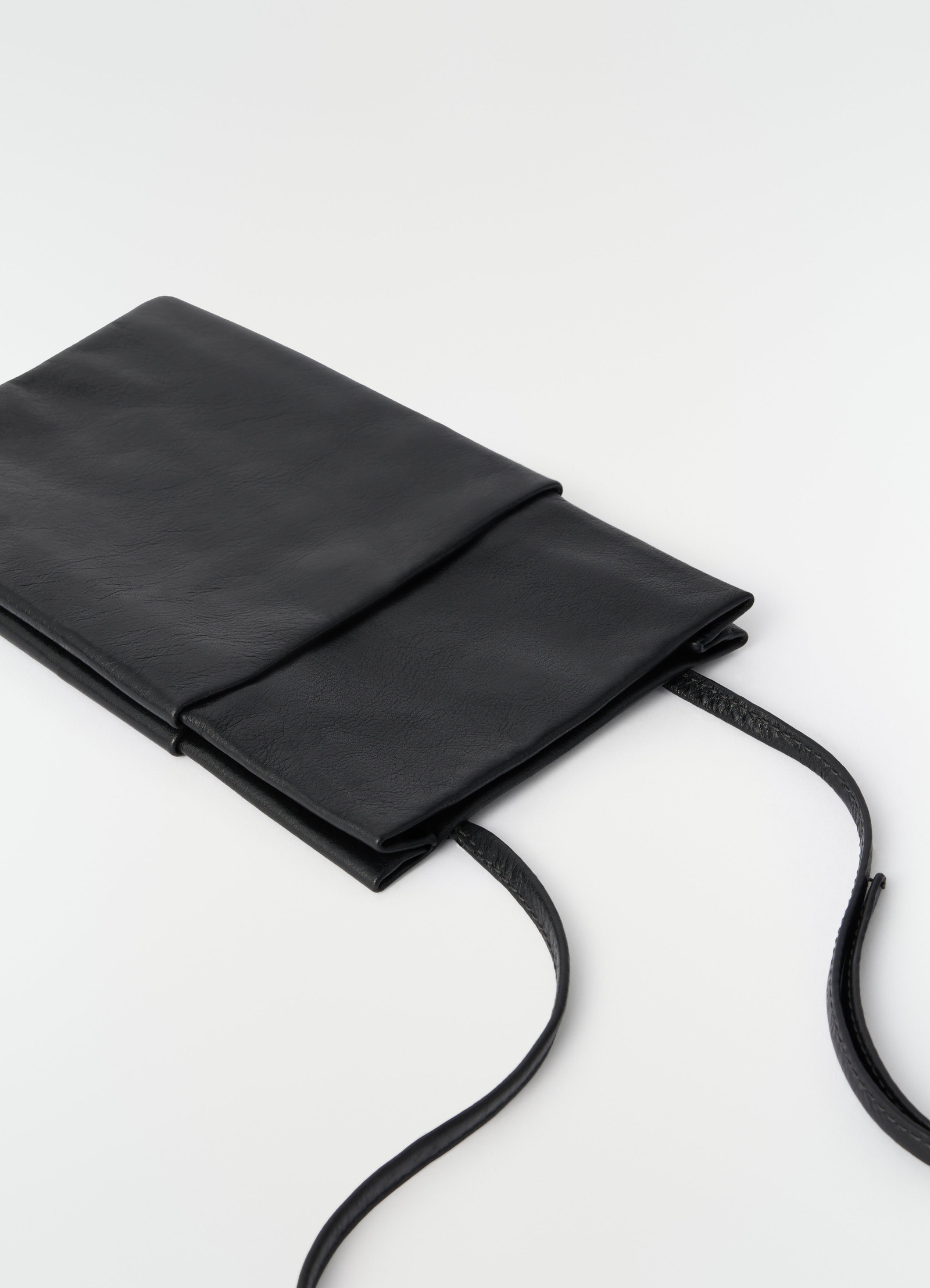 Book pouch in black grained leather by lemaire flatlay on white background with long strap visible